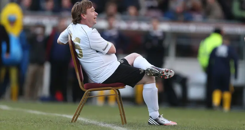 EastEnders legend Ian Beale sits on a chair in the middle of a football pitch